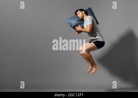 Junger schlafender Mann, der sein Kissen in einem leeren grauen Raum mit Kopierraum schweben hält Stockfoto