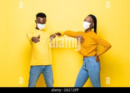 Das junge afroamerikanische Paar trägt Gesichtsmasken und stößt sich während der COVID-19-Pandemie in einem isolierten gelben Studiohintergrund an die Ellbogen, um sie zu begrüßen Stockfoto