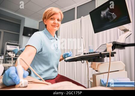 Sonografin, die den Patienten mit einem Ultraschallscanner untersucht. Arzt lächelt während der Durchführung von Ultraschalluntersuchungen in gynäkologischen Schrank. Konzept von Stockfoto