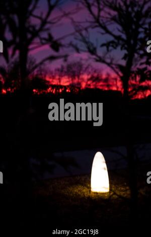 Verschwommene Nachtlichter im Park. Stimmungsvolle Lichtdeko im Garten mit Sonnenuntergang am Himmel. Laternenglühen in der Nacht. Atmosphärische Stimmung der goldenen Stunde. Stockfoto