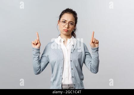 Skeptisches, enttäuschtes süßes asiatisches Mädchen empfehle dieses Produkt nicht, zeigend Finger oben mit störenden, beschwerlichen und düsteren Ausdrücken, haben Stockfoto