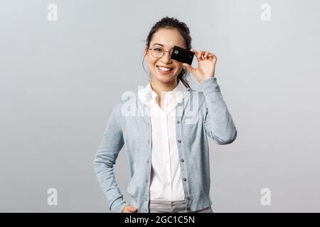 Technologie, Online-Käufe und Finanzierungskonzept. Gutaussehende, liebenswürdig asiatische Frau in Brille, die mit Kreditkarte über Augen zeigt und lächelt, empfehle die Bezahlung Stockfoto