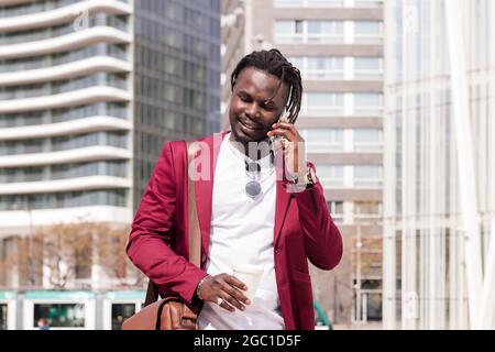 Eleganter afrikanischer Geschäftsmann geht durch die finanzielle Innenstadt der Stadt und telefoniert mit einem Kaffee in der Hand, einem Konzept der Technologie und einem Kommun Stockfoto