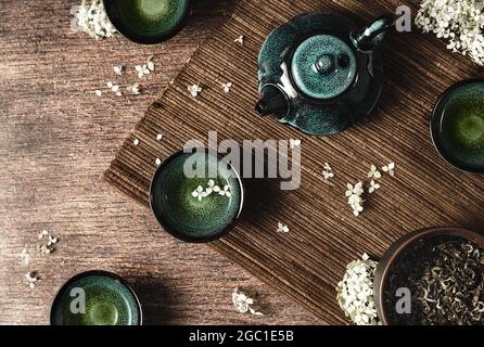 Dunkeltürkis glasierte Tonteapot und kleine Tassen auf dunklem Holzhintergrund mit Bambusmatte, verziert mit weißen Blumen. Kreativer minimalistischer Tee-Cerem Stockfoto