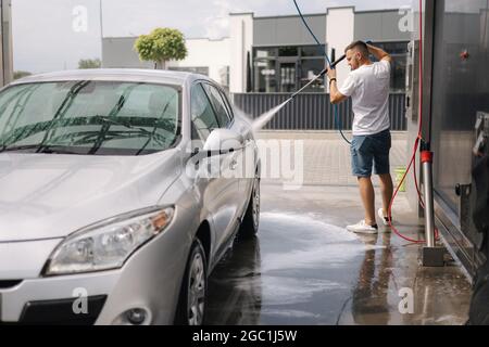 Junger Mann, der sein Auto mit einem Düsensprüher putzt. Autowäsche zur Selbstbedienung Stockfoto