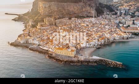 Sonnenaufgang über dem Hafen in Cefalu, Sizilien, Italien, Panorama-Luftaufnahme der Altstadt mit bunten Häusern am Wasser, Meer und La Rocca Klippe.attraktiv Stockfoto