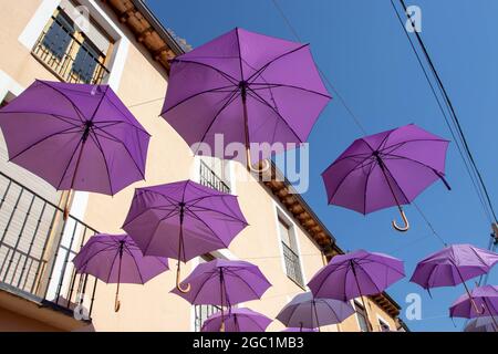 BRIHUEGA, SPANIEN - 10. JULI 2021: Lavendelfarbene Regenschirme, die während der blühenden Felder über der Straße hängen, Brihuega, Guadalajara, Castilla-La Stockfoto