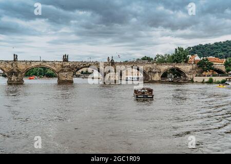 Prag, Tschechische Republik - August 4,2021. Kleine hölzerne Fähre Boot mit Passagieren überqueren Moldau, Karlsbrücke im Hintergrund. Lokalen Wassertransport Stockfoto