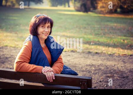 Eine kaukasische Frau mittleren Alters sitzt allein im goldenen Herbstpark. Reife Frau, Die Sich In Der Herbstlandschaft Entspannt Stockfoto