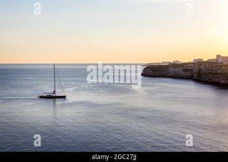 Ein Boot, das bei Sonnenuntergang auf einem ruhigen Meer in Korsika, Frankreich, in Richtung Küste segelt Stockfoto