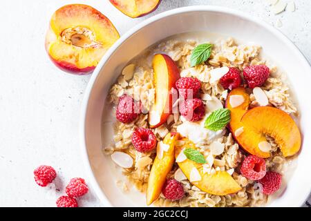 Sommer-Haferflocken mit Himbeeren, Pfirsichen und Nüssen in weißer Schüssel, Draufsicht. Gesundes vegetarisches Lebensmittelkonzept. Stockfoto