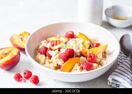 Sommer-Haferflocken mit Himbeeren, Pfirsichen und Nüssen in einer weißen Schüssel. Gesundes vegetarisches Lebensmittelkonzept. Stockfoto