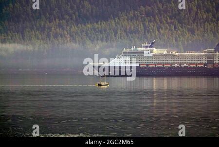 Alaska, Inside Passage, Pacific Coast; EIN riesiges Kreuzschiff stellt ein winziges Segelboot in den Schatten, während es sich langsam nach Norden bewegt, durch Nebel, der am frühen Morgen von Baranof Island entlang der Westküste Alaskas auf der Inside Passage aufsteigt. Stockfoto