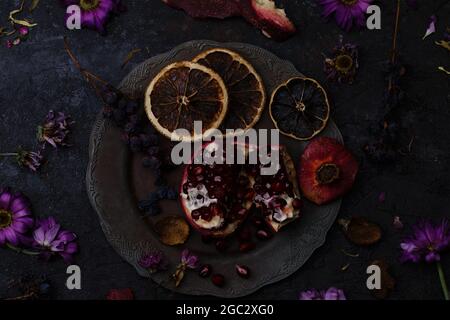 Reifer Granatapfel in einem Vintage-Teller, ein Bund trockener Trauben und orangefarbene Scheiben zwischen welkenden Blumen auf dunklem Steingrund Stockfoto