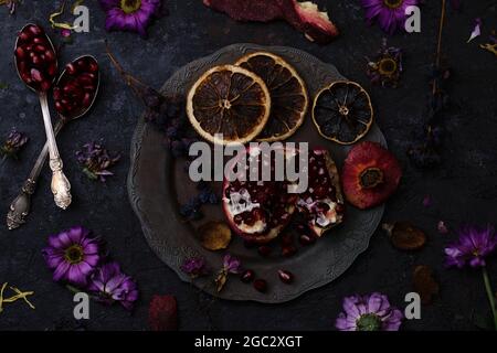 Geöffneter reifer Granatapfel auf einem Vintage-Teller, welke Blüten, Feijoa-Schale, trockene Früchte und Samen in Silberlöffeln Stockfoto