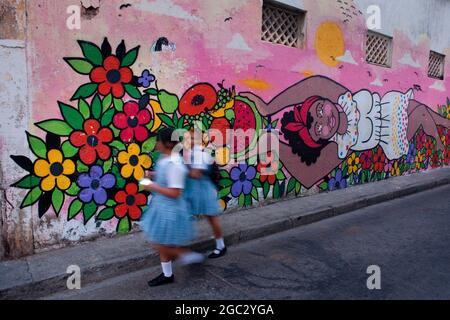 Lokale Schulkinder flanieren an einem farbenfrohen Wandgemälde mit Straßenkunst eines Palenquera-Obstverkäufers im Viertel Getsemani in Cartagena, Kolumbien. Stockfoto