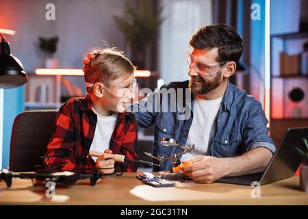 Positiver kaukasischer Vater lehrt seinen kleinen Sohn, wie man Displaykarten von einem modernen Laptop aus repariert. Mann und Junge arbeiten zu Hause mit Lötkolben. Stockfoto