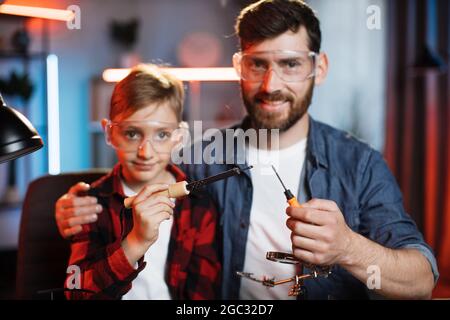 Positiver kaukasischer Vater umarmt seinen kleinen Sohn, während er zusammen am Tisch mit Schraubendreher und Lötkolben in den Händen sitzt. Zwei Männer, die Geräte reparieren, sind zu Hause. Stockfoto