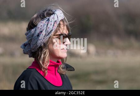 Eine attraktive Dame sieht mit einem Kopftuch lange Haare an windigen, aber sonnigen Tag trägt Sonnenbrille.Sie lächelt glücklich Stockfoto