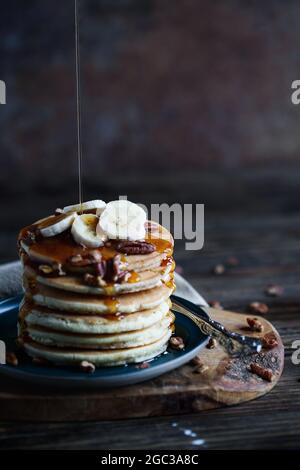 Ahornsirup, der auf Pfannkuchen mit frischen Bananenfrüchten und Nüssen gefüllt wird. Selektiver Fokus mit verschwommenem Hintergrund und Vordergrund. Stockfoto