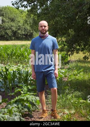 Ein junger weißer Kerl steht unter den Pflanzen. Ganzkörperfoto. Ruhiger, selbstbewusster Mann im Sommer im Garten. Stockfoto