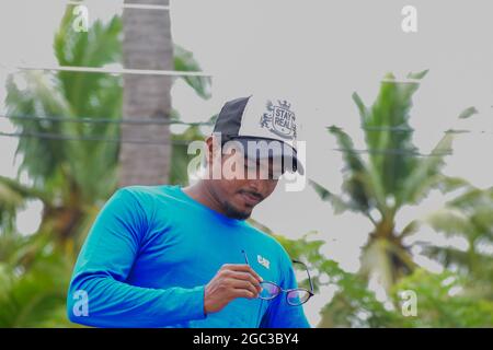 Junger indischer Mann in einem blauen Hemd und Baseballkappe betrachtet eine runde Brille im Freien. Stockfoto