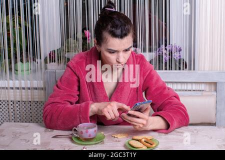 Portrait eines Mädchens in einem rosa Bademantel an einem Tisch schaut auf das Telefon, auf dem Tisch ist eine Tasse mit Tee und Frühstück. Telefon statt Frühstück Stockfoto