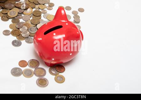 Viele Münzen verschiedener Länder liegen auf dem Tisch neben dem roten Sparschwein. Finanzkonzept. Stockfoto