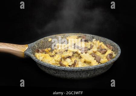 Gebratene Kartoffeln mit Pilzen in einer grau gefärbten Pfanne auf einem schwarzen Tisch, gerade gekocht, vom Braten dampfend, Nahaufnahme, dunkler Hintergrund Stockfoto