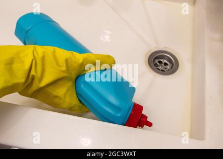 Eine Hand in einem gelben Handschuh gießt Waschmittel aus einer blauen Flasche auf ein Waschbecken. Das Konzept der Hausaufgaben, Desinfektion. Stockfoto