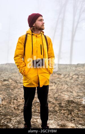 Junger männlicher Reisender in gelbem Mantel und Hut mit Kamera zum Entspannen im Freien bei nebligen Wetter. Urlaub und Lifestyle Wanderkonzept. Tourismus, Reisen, tr Stockfoto
