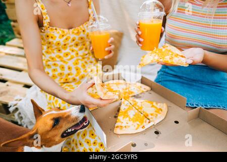 Zwei Frauen sitzen auf einem Sofa im Hinterhof und essen Pizza, trinken Limonade am sonnigen Sommertag. Stockfoto