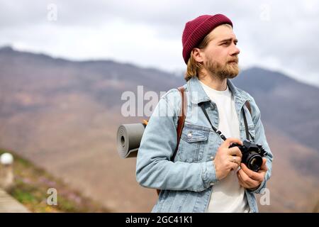 Reisender, der mit der Kamera in den Bergen unterwegs ist, die Landschaft genießt, Seitenansicht im Porträt. Bärtiger kaukasischer Rüde im lässigen Outfit im Freien, mit Rucksack. Stockfoto