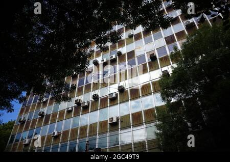 Altes Bürogebäude mit Rost an den Wänden und vielen Klimaanlagen an der Fassade des Gebäudes. Blick durch das üppige Laub Stockfoto