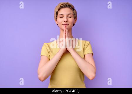Attraktive kaukasische Dame im lässigen T-Shirt beten, mit geschlossenen Augen halten die Hände zusammen, träumen, denken, mit einem netten Lächeln, isoliert auf purpl Stockfoto