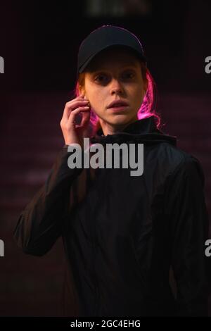 Porträt einer selbstbewussten Rothaarigen Frau in schwarzer Kappe und Kopfhörern, die allein in der Nachtstadt im Freien unterwegs ist, vor der Kamera posiert, Nachtlebenkonzept Stockfoto