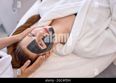 Junge kaukasische Frau nutzt die Dienste der professionellen Kosmetikerin bei at Spa. Cropped unkenntlich Meister Kosmetikerin setzt schwarze Maske auf Kunden fa Stockfoto