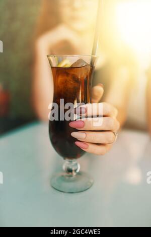 Frauenhand mit rosa und lila Maniküre halten Glas mit Cocktail. Das Mädchen in der Sommerbar genießt ein Eisgetränk. Lange lackierte Nägel. Helles Sonnenlicht. Stockfoto