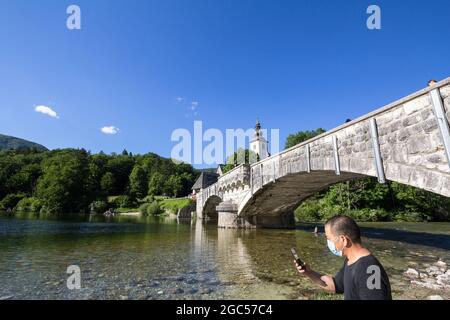 Bild der Kirche des heiligen johannes in Ribcev laz und ihrer ikonischen Brücke. Mit einem asiatischen Touristen, der sein Telefon benutzt, während er eine schützende Gesichtsmaske du trägt Stockfoto