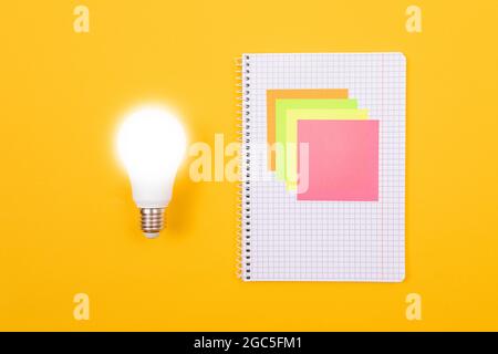 Ein Ideenkonzept - leuchtende Energiesparlampe und EIN Notizblatt mit Haftnotizen auf gelbem Tisch. Draufsicht, Flach Liegend, Leere Vorlage Stockfoto