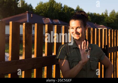 Glückliche Frau mit Hausschlüsseln in der Hand in der Nähe des Holzzauns ihres Hauses in einem Hüttendorf. Immobilienbesitzer, Makler, makler. Umzug, Kauf eines ho Stockfoto