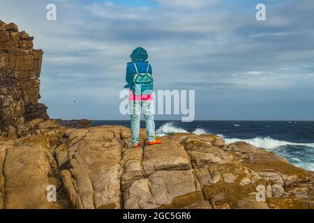 Eine Frau, die auf einem Felsen steht und das wilde Meer des Atlantischen Ozeans am Kap der Guten Hoffnung im Cape Point National Park, Südafrika, betrachtet. Stockfoto