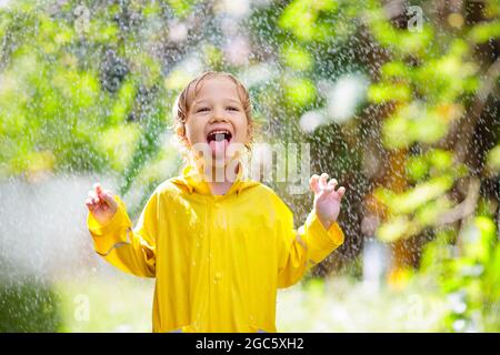 Kind spielt im Regen am sonnigen Herbsttag. Kind unter schwerer Dusche mit gelbem Entenschirm. Kleiner Junge mit wasserdichten Entenschuhen. Gummi Stockfoto