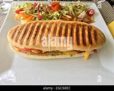 Ein Panini-Sandwich mit Schinken und Käse, serviert mit frischem Gemüsesalat auf der Seite, serviert auf einem weißen Teller. Stockfoto