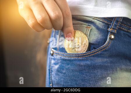 Nahaufnahme einer Frau, die Bitcoin in die Jeanstasche steckte, goldene Kryptowährung-Münze im Taschenhintergrund Stockfoto