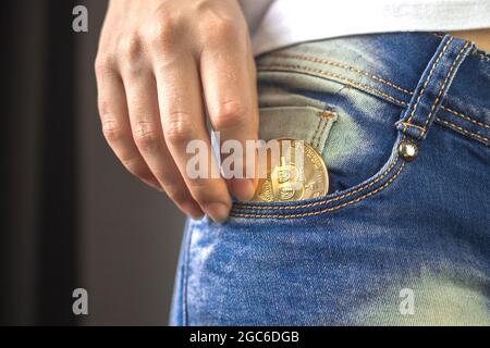 Frau Hand setzen Bitcoin in Jeans Tasche, goldene Kryptowährung Münze in Tasche Hintergrund Stockfoto