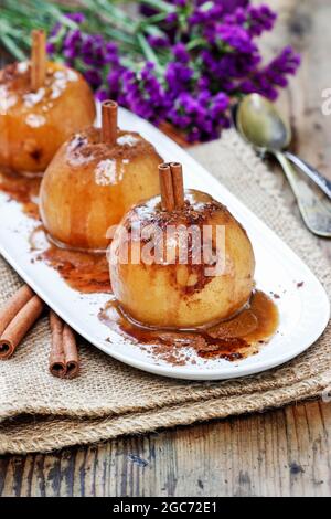 Äpfel in Karamell. Teller steht auf einem schönen Holztisch, violette Dekorationen und Duftkerzen im Hintergrund. Stockfoto