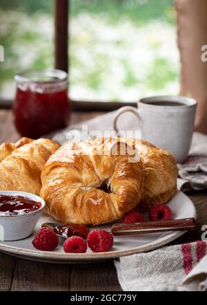 Croissants mit Himbeerkonfitüre und frischen Himbeeren auf einem Teller am Fenster Stockfoto