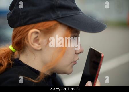 Das Mädchen nimmt auf der Straße eine Sprachnachricht auf. Die Frau kommuniziert auf einem Smartphone. Der Teenager spricht in den Telefonbildschirm. Frau in einer Mütze. Stockfoto