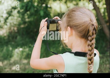 Ein glückliches kleines sieben Jahre altes Mädchen fotografiert eine Sommer-Naturlandschaft mit einer Kamera mit Live-Ansicht. Kinder adoptieren ihre Eltern Hobbys. Summe Stockfoto